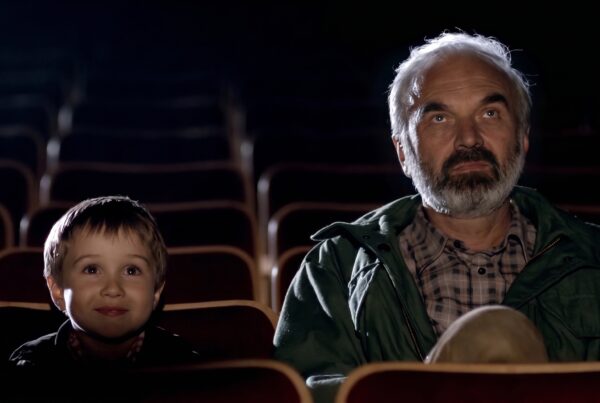 Louka and the young Kolya sit a few seats apart in a darkened cinema auditorium, their faces lit by the light of the screen as they watch.