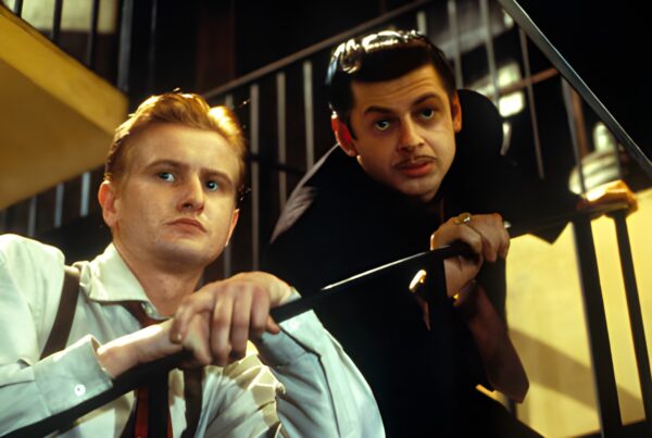 Two men in shirtsleeves and braces lean on a stair rail inside the club, one fair-haired and one dark with a moustache, both looking off to one side.